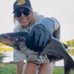 Woman holding a large fish by the water, smiling.