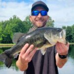 Man proudly holding a large bass fish by a lake.