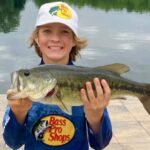 A smiling boy holding a largemouth bass by a lake.