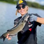 Woman proudly holding a large fish by the water.