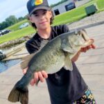 Young angler proudly holds a large bass fish by the water.