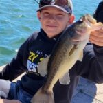 A boy proudly holding a largemouth bass by the water.