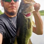 Man holding a large bass fish near a lake.