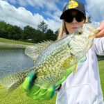 Person holding a large colorful fish near a lake with a blue sky.