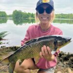 A woman proudly holding a large fish by the lakeside.