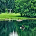 A peaceful lake with two people fishing and a small pavilion in the background.