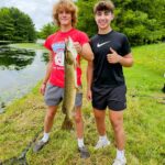 Two young men proudly display a large fish by a lakeside.