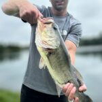 Man holding a large bass fish caught by a lake.