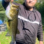 Man holding a large bass fish outdoors in sunlight.
