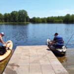 Two people fishing from small boats near a dock on a calm lake.