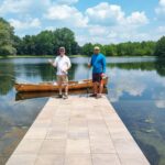 Two people standing on a dock with a canoe by a calm lake.