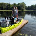 Two men standing on a dock with fishing gear and kayaks by a lake.