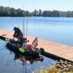Person preparing fishing gear on a kayak by a calm lake dock.