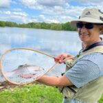 Smiling man holding a freshly caught fish by a lake.