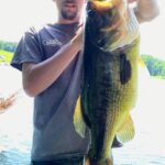 Man holding a large bass fish near a lake.