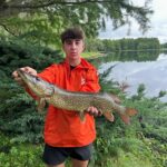 A young boy holding a large fish by a lake.