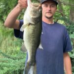 Man proudly holds a large bass fish in a lush green outdoor setting.