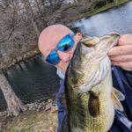 Man wearing blue sunglasses holding a large fish near a river.
