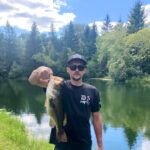 Man proudly holds a large fish by a scenic lake.