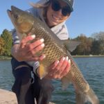 Person holding a large fish by a lake on a sunny day.