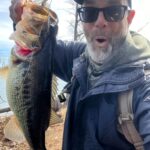Man excitedly holds a large fish by the water.