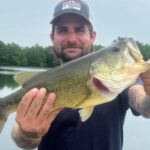 Man proudly holding a large fish he caught near a lake.