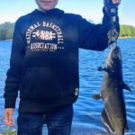 Person proudly holding a large catfish by the water.