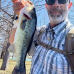 Man proudly holding a large bass fish by the water.