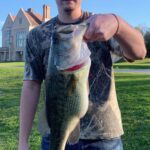 A person holding a large bass fish outdoors near a house.