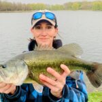 Woman proudly holding a large fish by a lake.