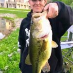 Person proudly holding a large bass fish caught by a pond.