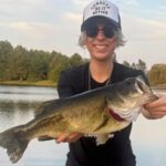 Person proudly holding a large fish by a lake at sunset.