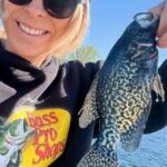 Smiling woman holding a large crappie fish near a lake on a sunny day.