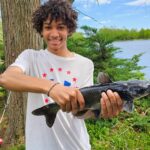 A young boy proudly holding a freshly caught fish near a lake.