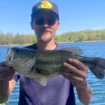 Man holding a large fish by a lake on a sunny day.