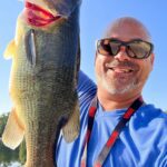 Man smiling, holding a large fish he caught outdoors.