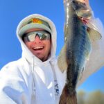 Person in white hoodie holding a large fish, smiling under a clear blue sky.