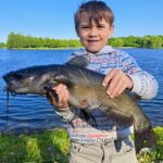 Young boy proudly holding a large catfish by a lake.